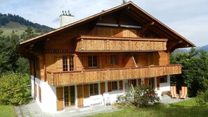 Een chalet van hout met twee verdiepingen en een balkon, omringd door groen en bergen.