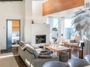 A modern living room with a sofa, coffee table, and chairs. There is a TV, a lamp, and a window with curtains.