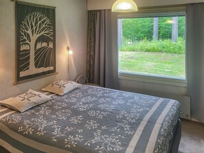 A bedroom with a bed, a wall-mounted lamp, a window with a view of greenery, and a painting of a tree.