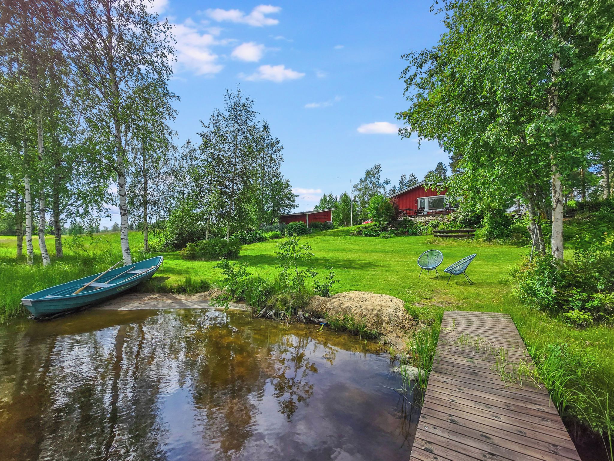 Eine ruhige Seenlandschaft mit einem hölzernen Steg, einem kleinen Boot und einem roten Gebäude mit Terrasse.