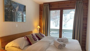 Ein ordentlich bezogenes Bett mit weißen und roten Kissen, ein hölzernes Kopfteil und ein Blick auf eine verschneite Landschaft durch ein großes Fenster mit Vorhängen.