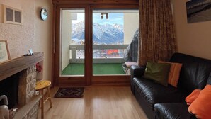 Ein gemütliches Wohnzimmer mit Kamin, einer schwarzen Ledersofa und Blick auf schneebedeckte Berge durch eine Schiebetür.