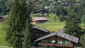 A traditional chalet-style building with a balcony, surrounded by lush greenery and other similar buildings in a mountainous area. - Les Silenes by Interhome (Gstaad)
