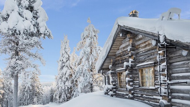 Eine Blockhütte mit schneebedecktem Dach und Fenstern, umgeben von schneelastigen Bäumen.