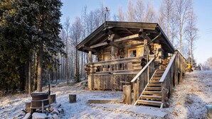 A log cabin with a wooden staircase leading to a porch, surrounded by a snowy forest.