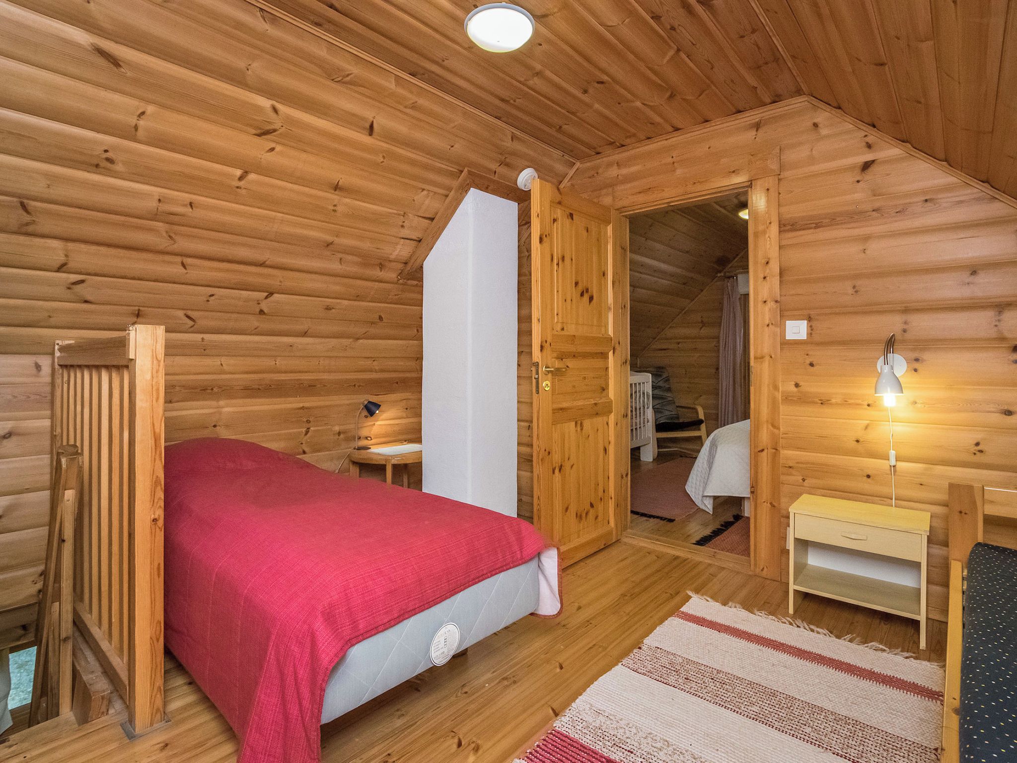A wooden cabin bedroom with a bed, a nightstand, a lamp, and a door leading to another room.