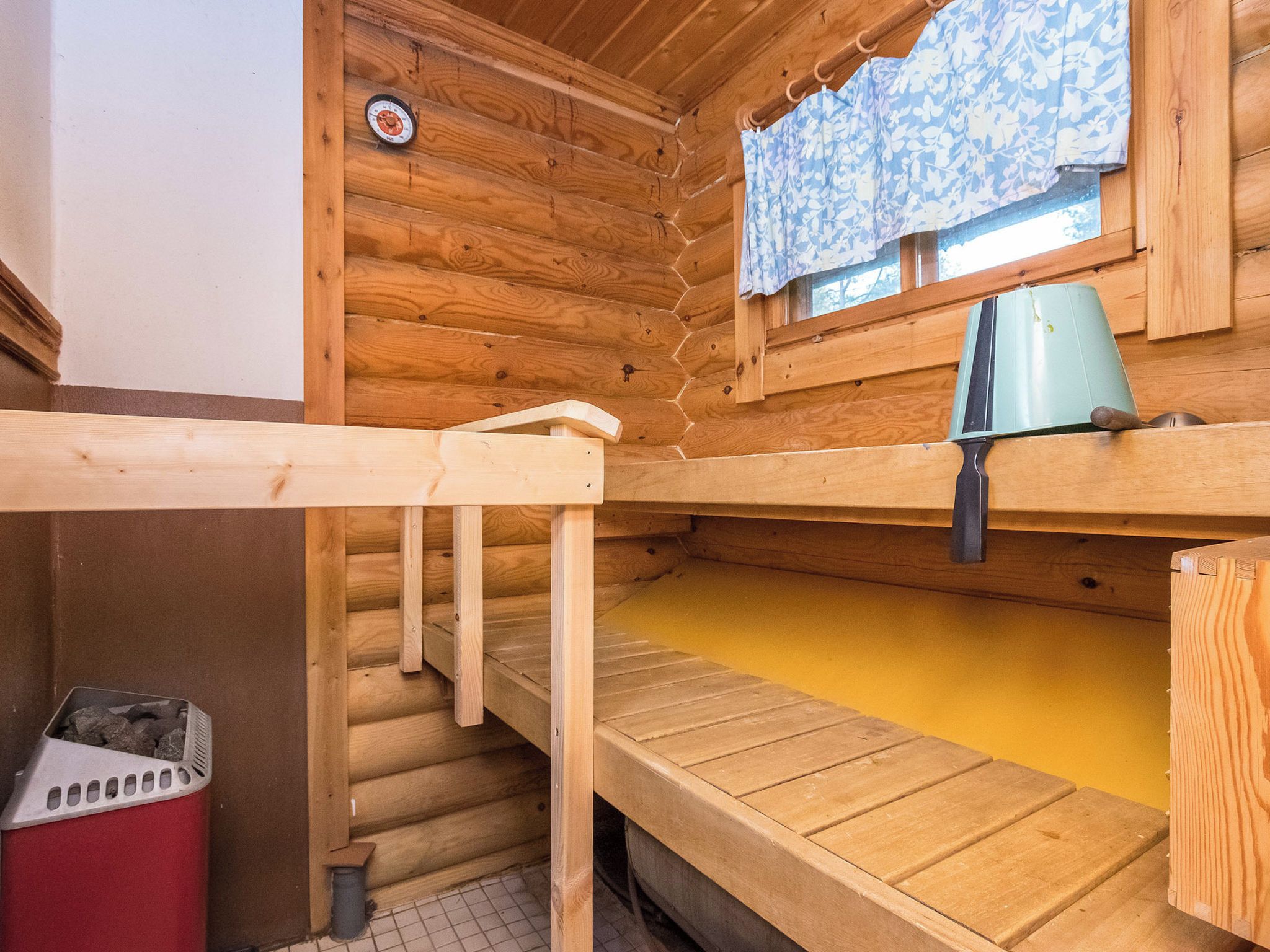 A wooden cabin interior with bunk beds, a window with a floral curtain, and a small heater.