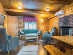 A cozy cabin interior with a wooden ceiling, a sofa, two armchairs, a dining table, and a fireplace.