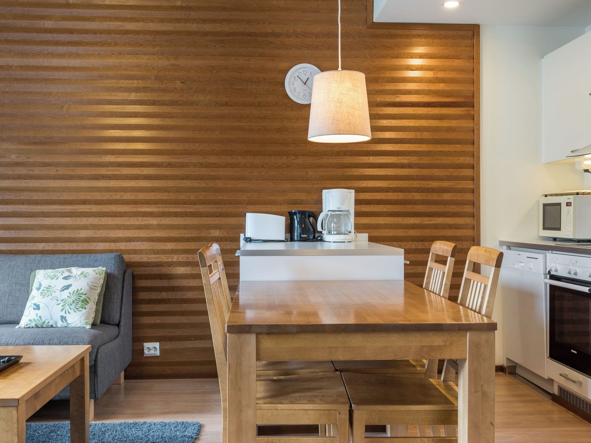 A modern kitchen with a wooden wall, a dining table with chairs, a coffee maker, and a microwave.