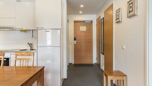 Eine kleine Küche mit einem Kühlschrank, einem Esstisch und Stühlen. Eine Holztür führt zu einem Flur.