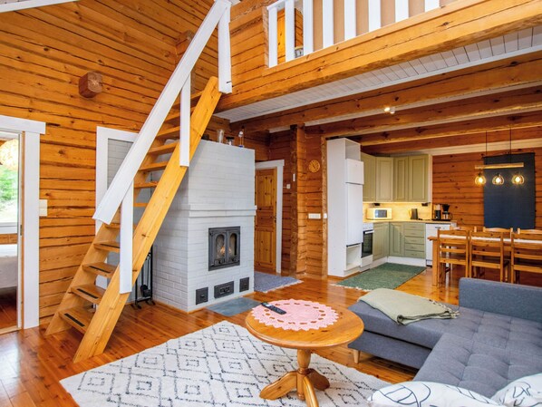 A wooden cabin interior with a fireplace, staircase, kitchen, and seating area.