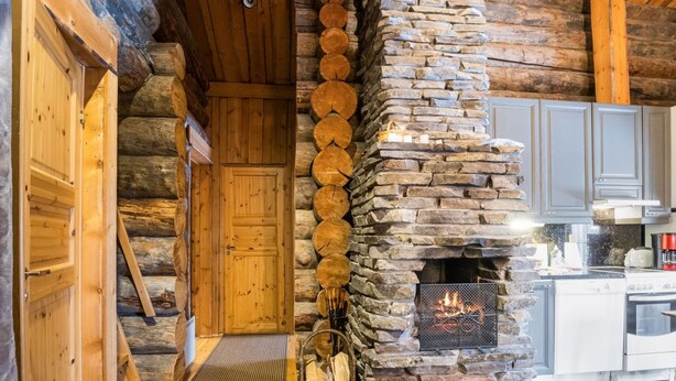 Interior de una cabaña de troncos con chimenea de piedra, puerta de madera y zona de cocina.