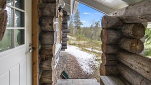 Una cabaña de troncos con puerta y ventanas de madera, un porche con vistas a un paisaje nevado y árboles.
