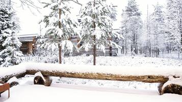 A log bench in a snowy forest with a log cabin in the background.