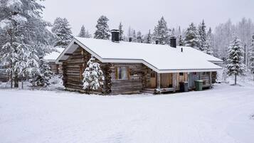 A log cabin with a snow-covered roof and a chimney, surrounded by snow-laden trees.