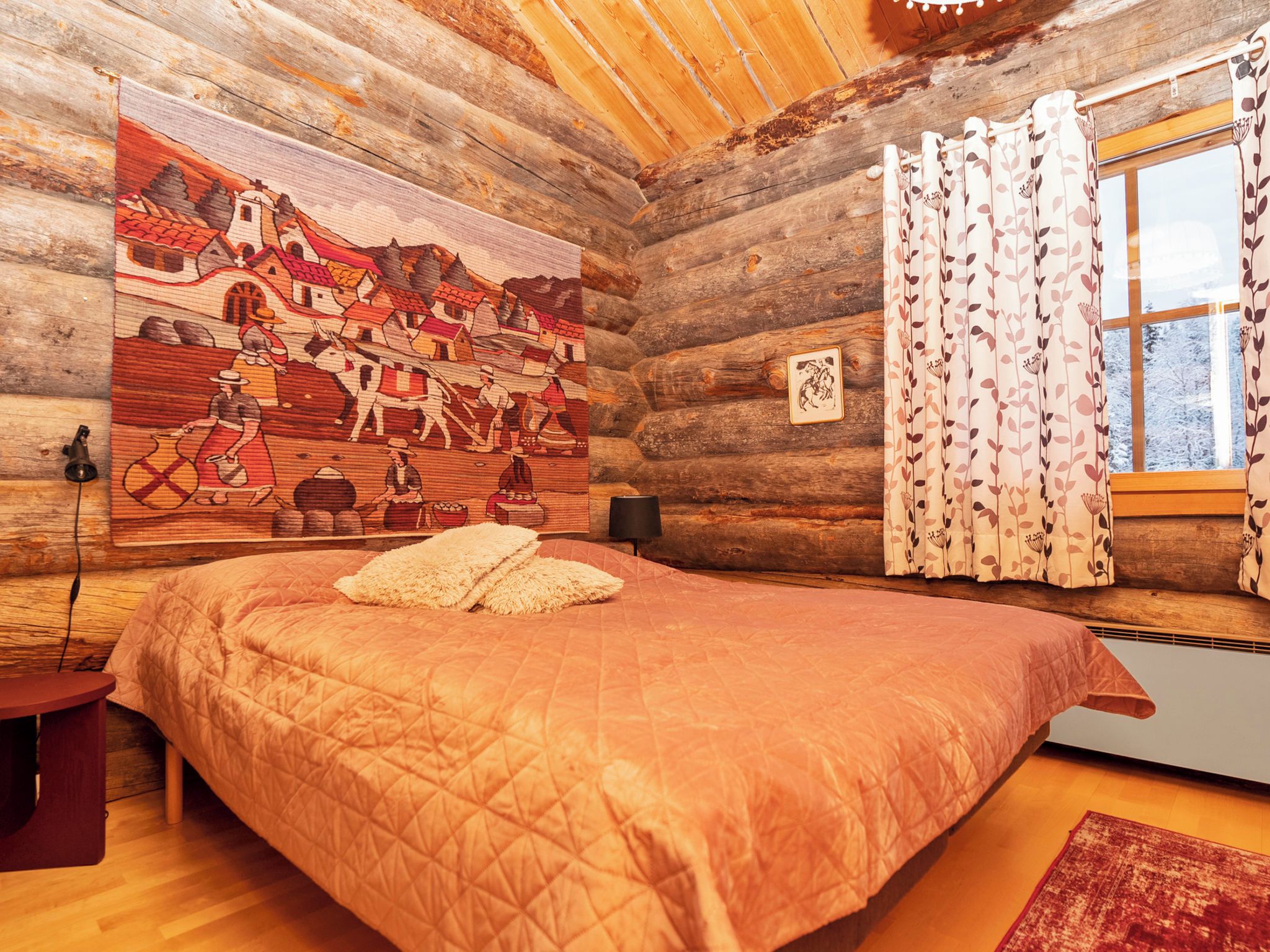 A log cabin bedroom with a large bed, a wooden wall with a mural, a window with curtains, and a small bedside table.