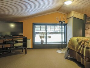 Een kamer met een televisie op een standaard, een raam met zonwering, een kamerplant en een bed met een dekbed.