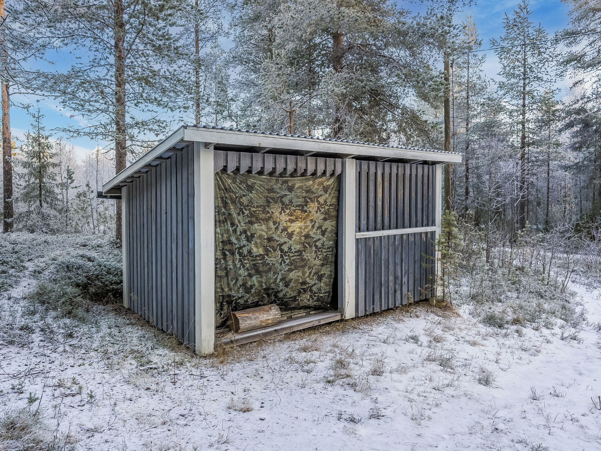 Una piccola baita di legno con una porta a fantasia camouflage, immersa in un bosco innevato.