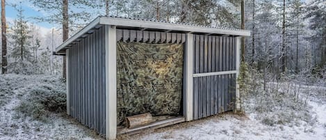 Una pequeña cabaña de madera con una puerta de estampado camuflado, rodeada por un bosque nevado.