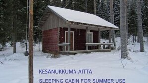 Une petite cabane rustique avec un porche, entourée de neige et d’arbres, et indiquée comme étant une cabane servant de lieu de couchage pour l’été.