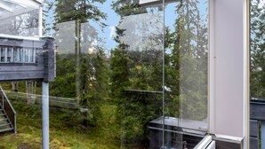 A modern building with large glass windows, a wooden deck, and a view of a forested area.