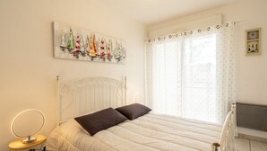 A bedroom with a bed, a bedside table with a lamp, a radiator, and a window with curtains. - Le Forum by Interhome (Royan)