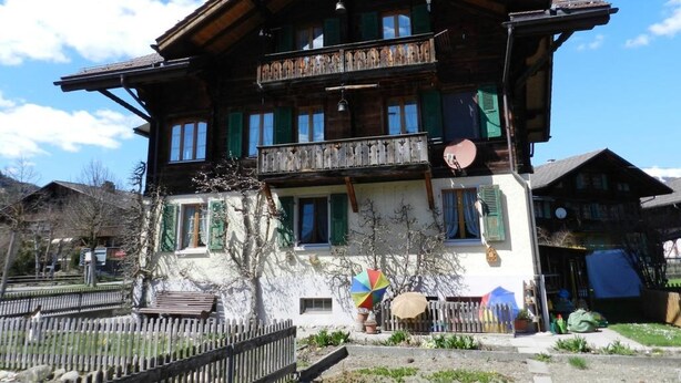 Un chalet traditionnel en bois, doté d’un balcon, d’un petit jardin et surplombant un ciel d’un bleu éclatant.