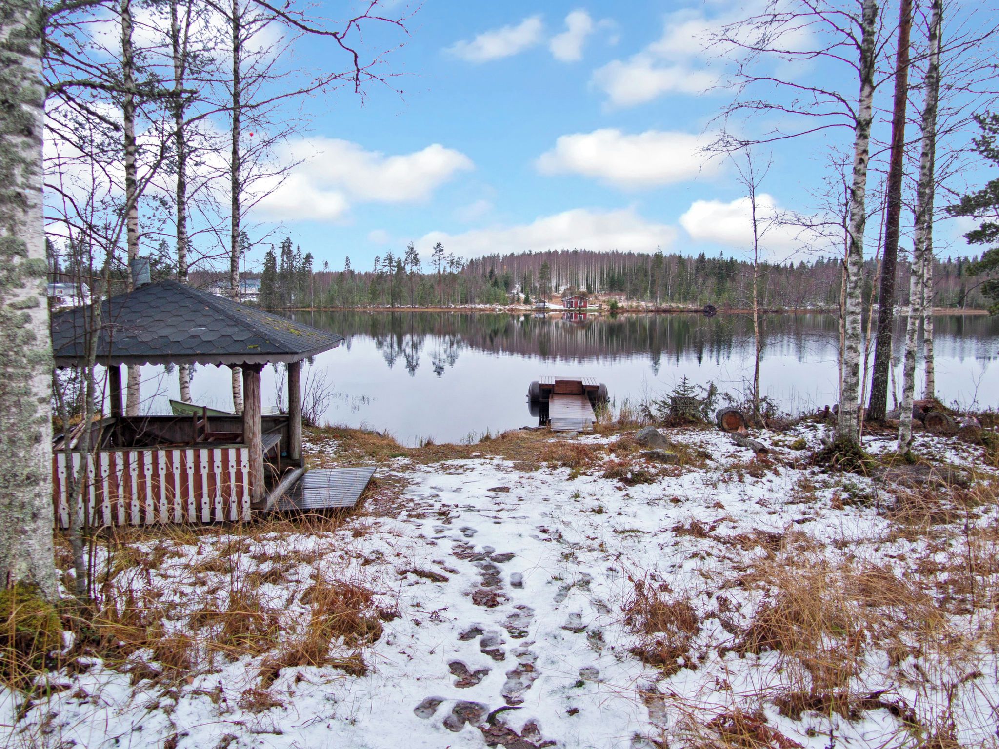 Mökki, jonka edessä on laituri, joka ulottuu tyynen järven rantaan. Mökkiä ympäröivät puut, ja maassa on lunta.
