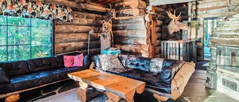L’intérieur d’un chalet rustique, avec une cheminée surmontée de têtes d’animaux empaillées, un canapé en cuir noir et une table basse en bois.