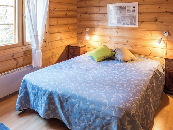 A wooden room with a bed, a nightstand, a wall-mounted light, and a window with curtains.