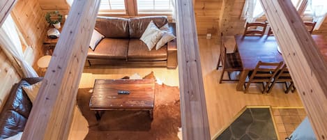 Ein Interieur einer Hütte mit Holzbalkendecke, einem braunen Ledersofa, einem Holzesstisch und einem Steinfußboden.
