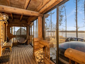Une cabane en bois avec une terrasse en bois, un fauteuil suspendu et une vue sur une voiture et des arbres à l’extérieur.