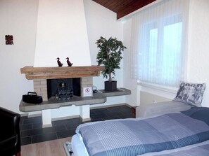 Un dormitorio con chimenea, una cama, un ventanal con cortinas ligeras y una planta en maceta.