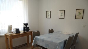 Un pequeño comedor con una mesa, sillas, una cafetera y un ventanal con cortinas ligeras.