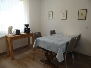A small dining area with a table, chairs, a coffee maker, and a window with sheer curtains.