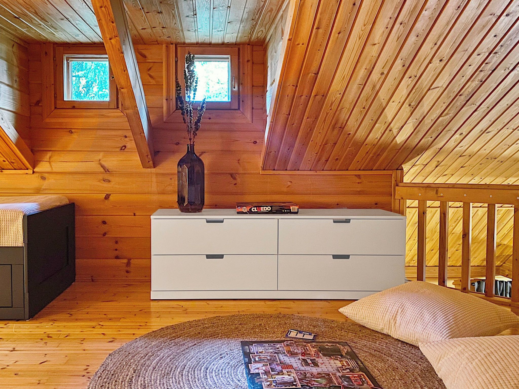 A cabin interior with a loft bedroom, wooden walls, a white dresser, a window with a view, and a woven rug.