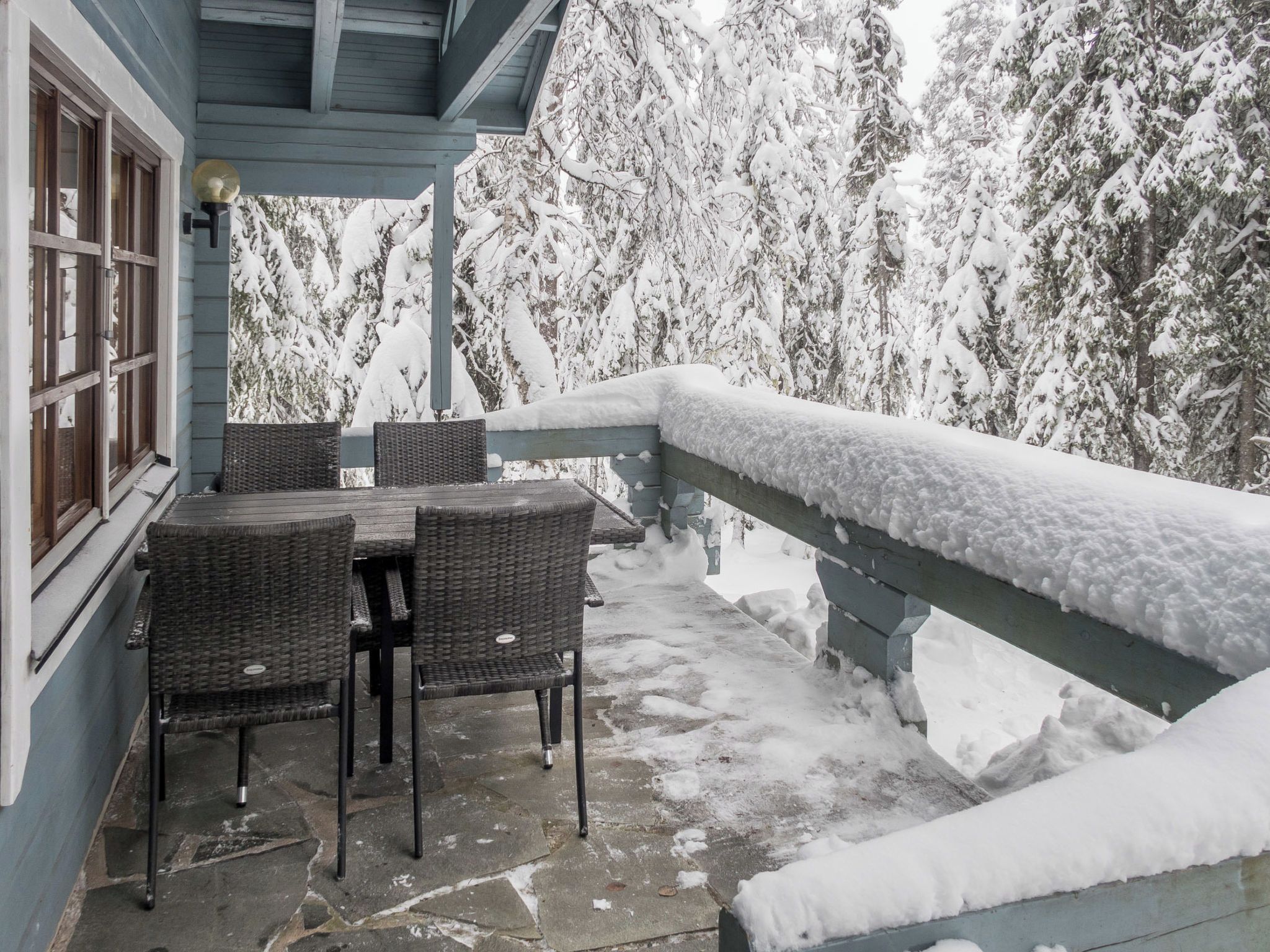 A covered outdoor seating area with wicker chairs and a table, surrounded by snow-covered trees.