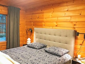 A bedroom with a wooden ceiling, a bed with patterned bedding, a bedside lamp, and a window with curtains.