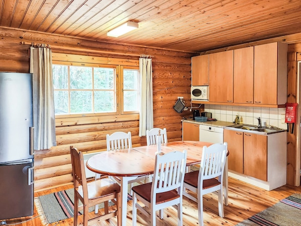 A cabin interior with a wooden dining table, chairs, a kitchen with wooden cabinets, a refrigerator, and a window with curtains.