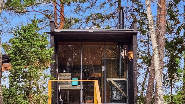 Une cabane dotée d’une terrasse en bois, d’une cheminée et d’une fenêtre donnant sur une forêt.