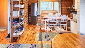 L’intérieur chaleureux d’un chalet, avec un revêtement mural en bois, un coin repas avec des chaises blanches, un réfrigérateur et une fenêtre avec des rideaux.