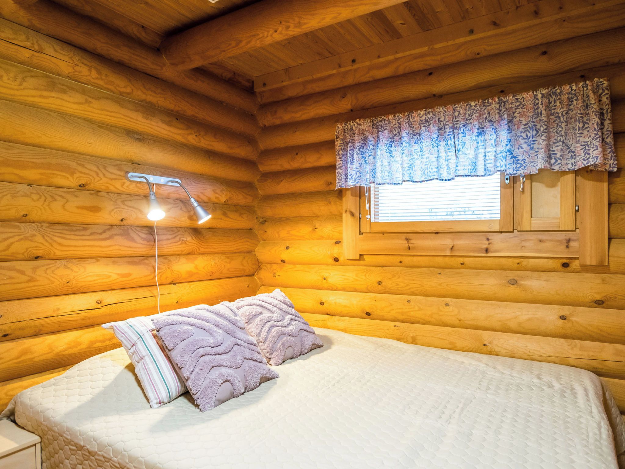 Un dormitorio de cabaña de troncos con una cama, dos almohadas, una mesita de noche, una lámpara y una ventana con cortina floral.