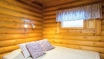 Un dormitorio de cabaña de troncos con una cama, dos almohadas, una mesita de noche, una lámpara y una ventana con cortina floral.
