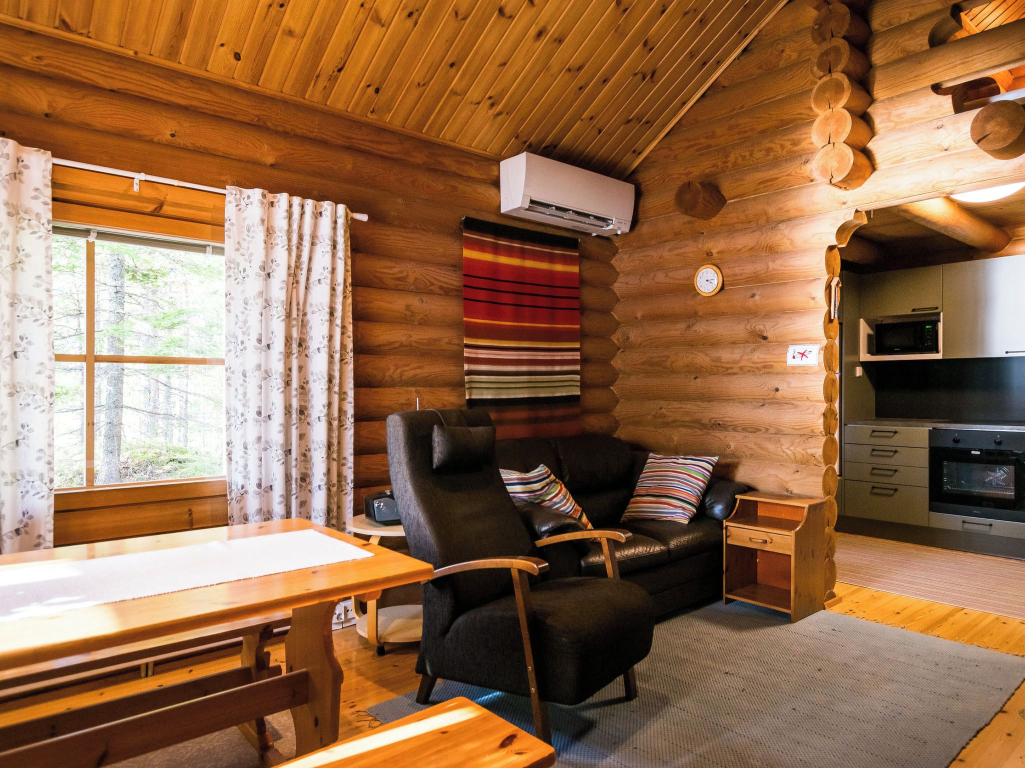 Interior de una cabaña de troncos con una mesa de comedor de madera, un sofá de cuero con cojines, una pequeña mesita auxiliar y una zona de cocina con microondas y horno.