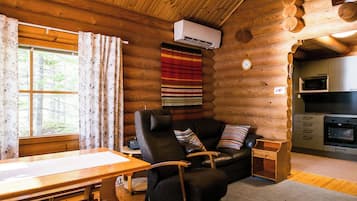 Interior de una cabaña de troncos con una mesa de comedor de madera, un sofá de cuero con cojines, una pequeña mesita auxiliar y una zona de cocina con microondas y horno.