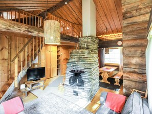 A log cabin interior with a stone fireplace, wooden staircase, and a television on a wooden stand.