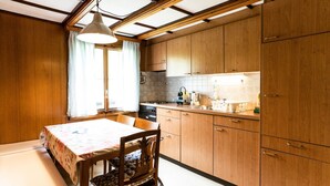 A compact kitchen with wooden cabinets, a dining table with a floral tablecloth, and a single chair. - Bürcher Haus 1. OG Süd-West by Interhome (Riederalp)