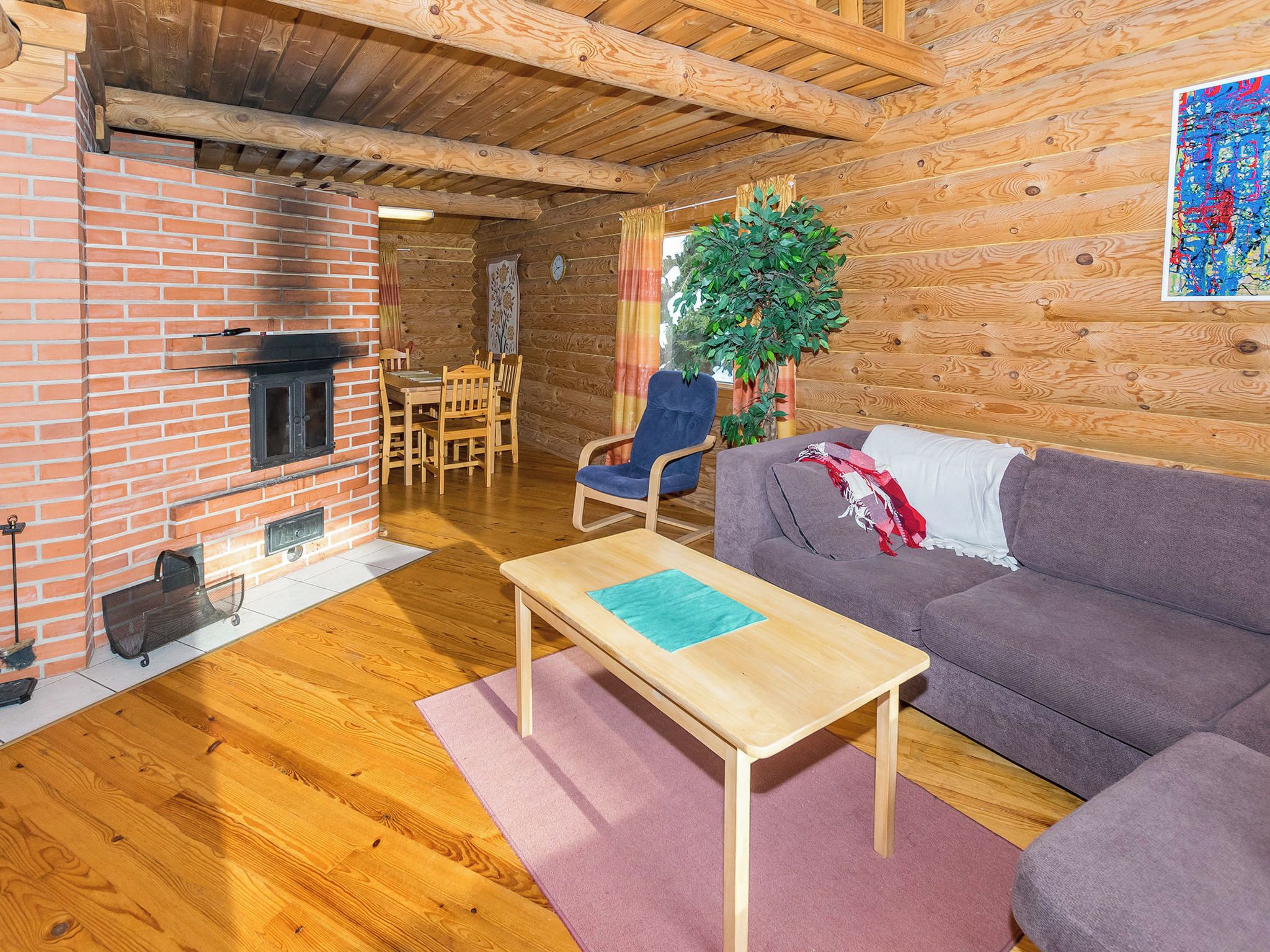 A cozy living room with a brick fireplace, wooden furniture, and a large plant.