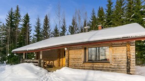 Una cabaña de troncos con techo nevado, un porche de madera y una ventana.