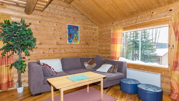 Tunnelmallinen mökin sisustus, jossa on puinen hirsiseinä, harmaa sohva, puinen sohvapöytä ja suuri ikkuna, jossa on verhot.